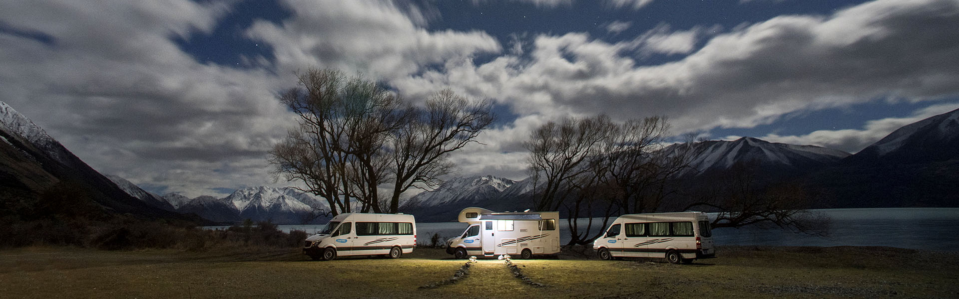 3 motorhomes sit head to tail along the edge of a lake as night falls around them